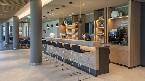 A modern bar area with a long counter, bar stools, and shelves stocked with bottles and glasses. - Holiday Inn Express Düsseldorf – Hauptbahnhof by IHG (Düsseldorf)