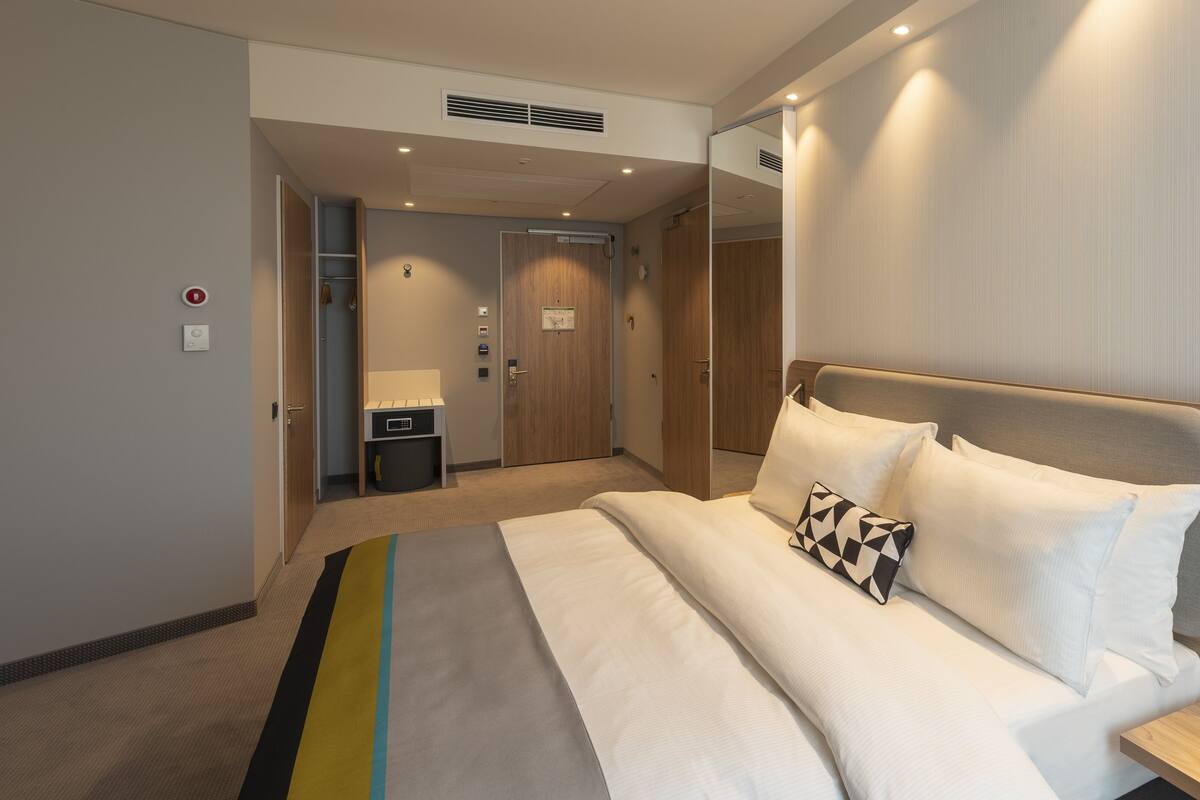 Ein modernes Hotelzimmer mit einem ordentlich bezogenen Bett, einem Nachttisch mit Lampe, einem Spiegel und einem kleinen Schreibtisch mit Stuhl.
