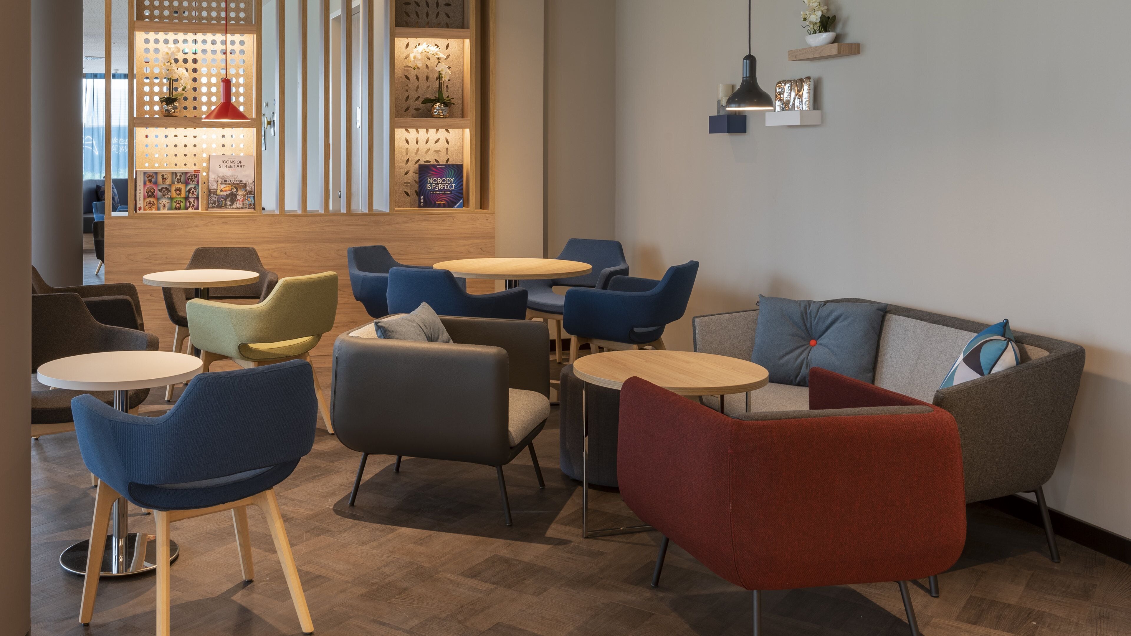 A modern lounge area with multiple seating options including sofas and chairs, wooden tables, and a decorative wall with perforated panels and lighting.