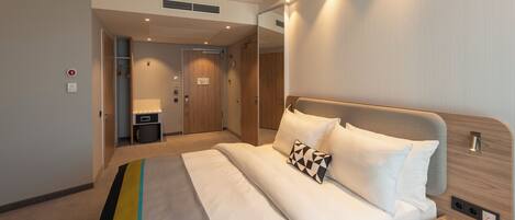 Habitación de hotel moderna con una cama bien hecha, una mesita de noche con lámpara, un espejo y un pequeño escritorio con silla.