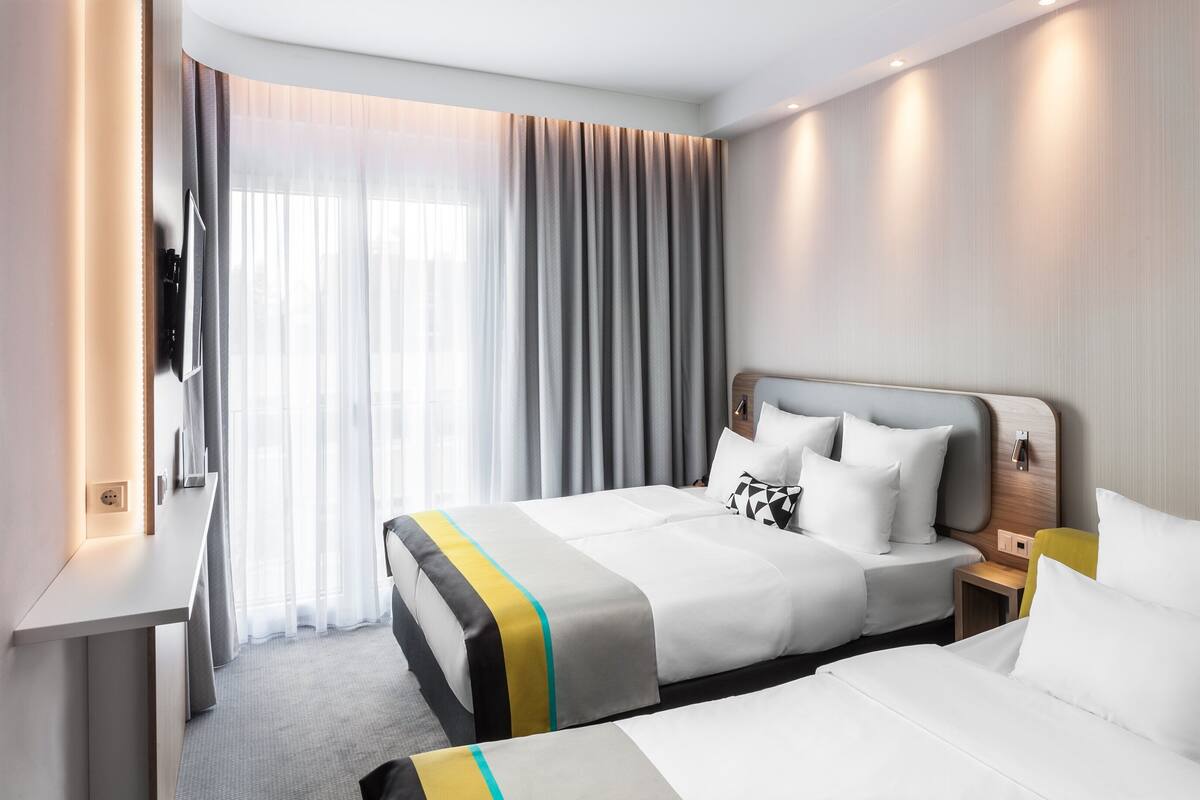 Ein Hotelzimmer mit zwei Betten, einem Schreibtisch und einem großen Fenster mit transparenten Vorhängen.