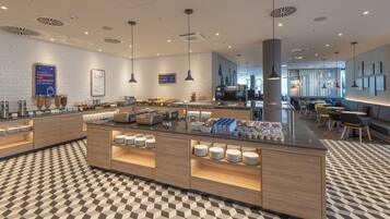 Un moderno hall de hotel con zona de buffet central, arreglos de asientos y mostrador de recepción.