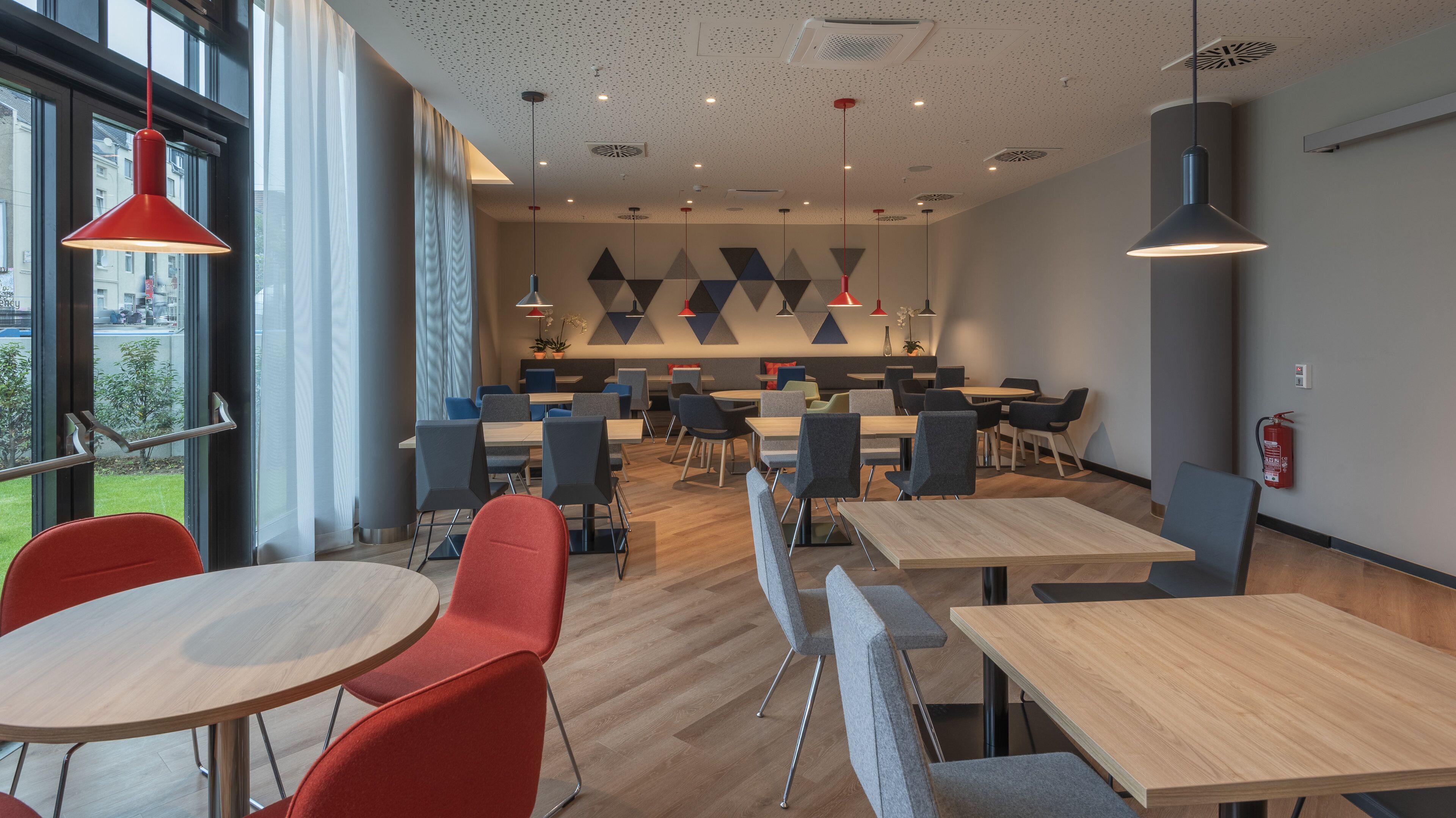 A modern dining area with round tables and chairs, a fire extinguisher on the wall, and a view of the outside through large windows.