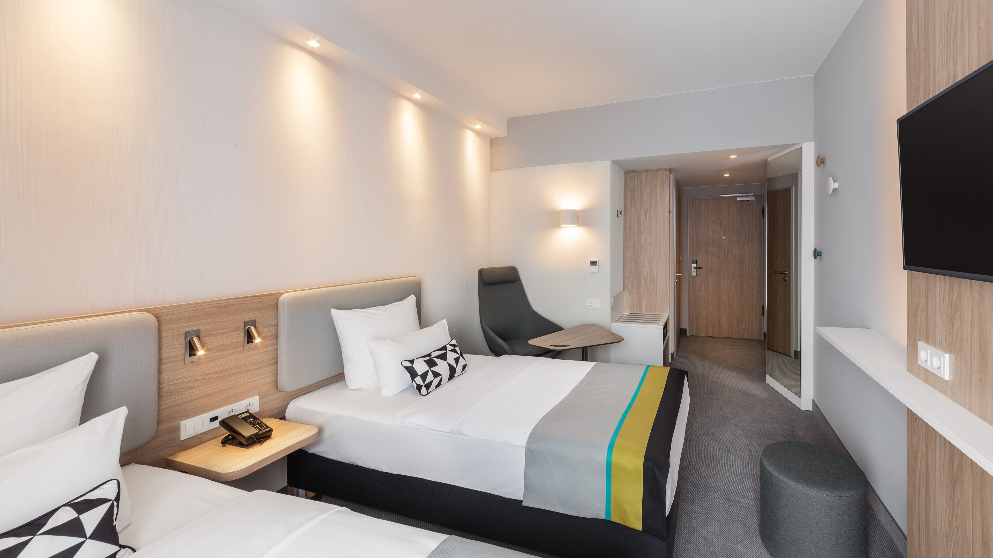 A modern hotel room with a bed, bedside tables, a chair, a desk, and a TV.