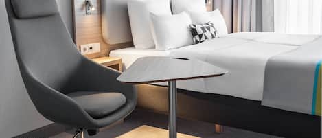 A modern hotel room with a grey upholstered armchair, a wooden nightstand, a bed with white bedding, and a small table with a grey tabletop.