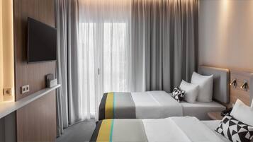 Habitación de hotel con televisión montada en la pared, cama con colcha a rayas, mesita de noche con lámpara y ventana con cortinas traslúcidas.