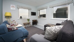 Een woonkamer met een bank, een salontafel en een aan de muur bevestigde televisie. Er zijn ramen met behang en uitzicht op buiten.