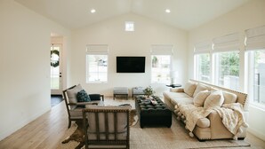 A spacious living room with a high ceiling, a flat-screen TV mounted on the wall, a beige sofa, a wooden chair, and a large window with blinds. - Luxury in DT Tempe -2 en-suites - ASU, Spring Training, Golf & More (Tempe)