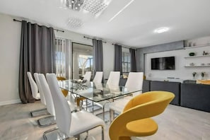 Een moderne eetruimte met een glazen tafel en witte stoelen, een gele fauteuil en een grijze bank op de achtergrond.