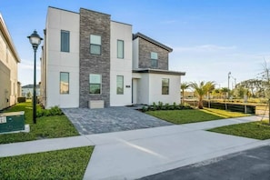 A modern two-story residential building with a flat roof, large windows, and a paved driveway.