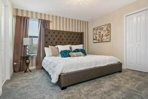 A bedroom with a tufted headboard, a bed with white bedding and teal pillows, a bedside table with a lamp, a window with curtains, and a door.