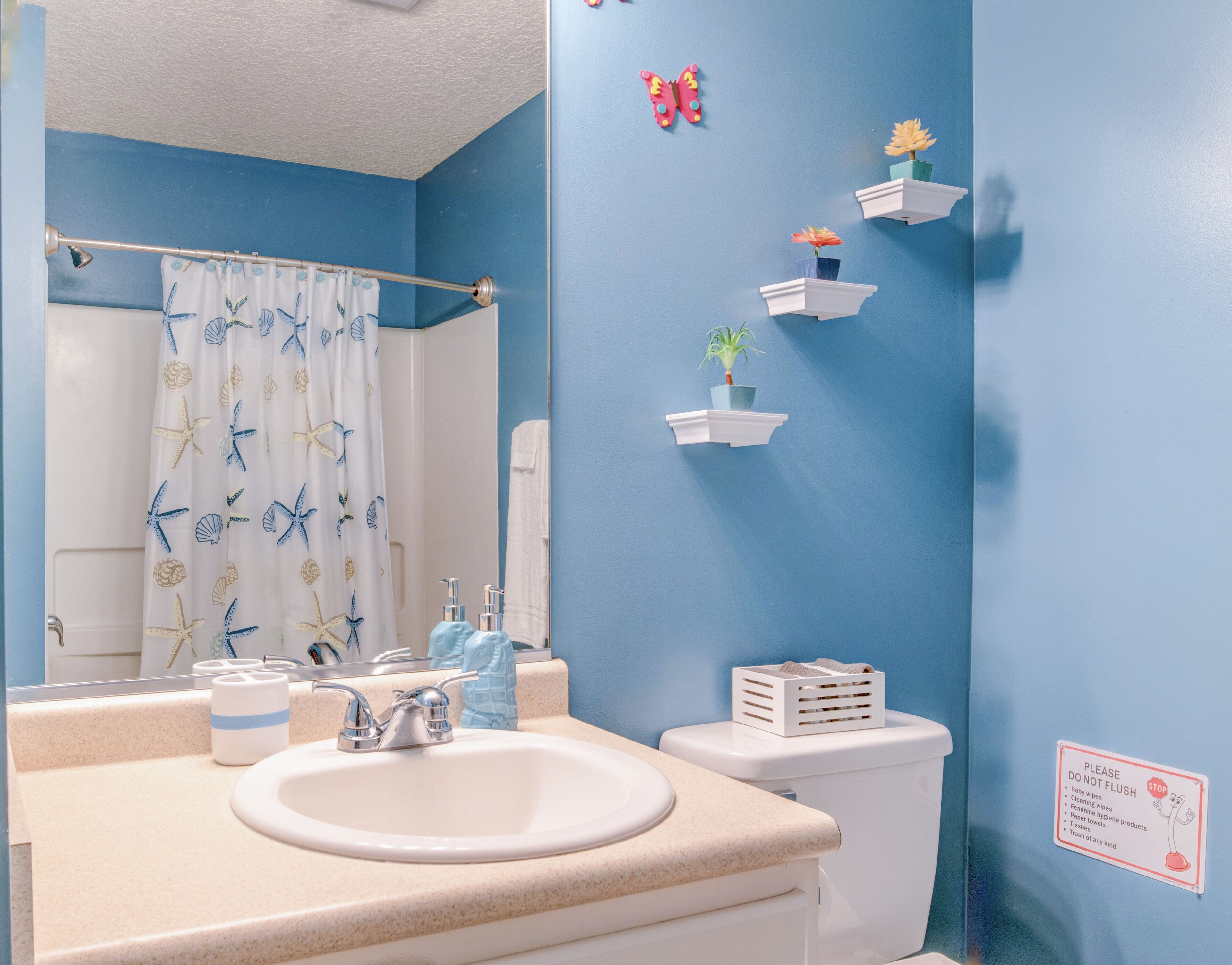 A bathroom with a blue wall, a white sink, a toilet, and a shower curtain with starfish and seashells. There are also small shelves with plants and a sign on the wall.