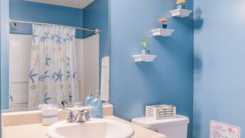 A bathroom with a blue wall, a white sink, a toilet, and a shower curtain with starfish and seashells. There are also small shelves with plants and a sign on the wall.