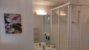 Een badkamer met een wit wastafel, spiegel en douche. Op de muur hangt een lijst met bloemen.