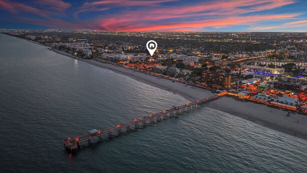 An aerial view of a coastal city with a long pier extending into the sea, a beach, and a densely populated urban area with illuminated buildings.