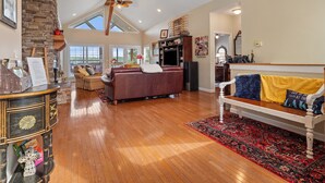 A spacious living room with a wooden floor, a stone fireplace, a large window, and a ceiling fan. - Blue Door - Entertainment area downstairs with a pool table (Arley)