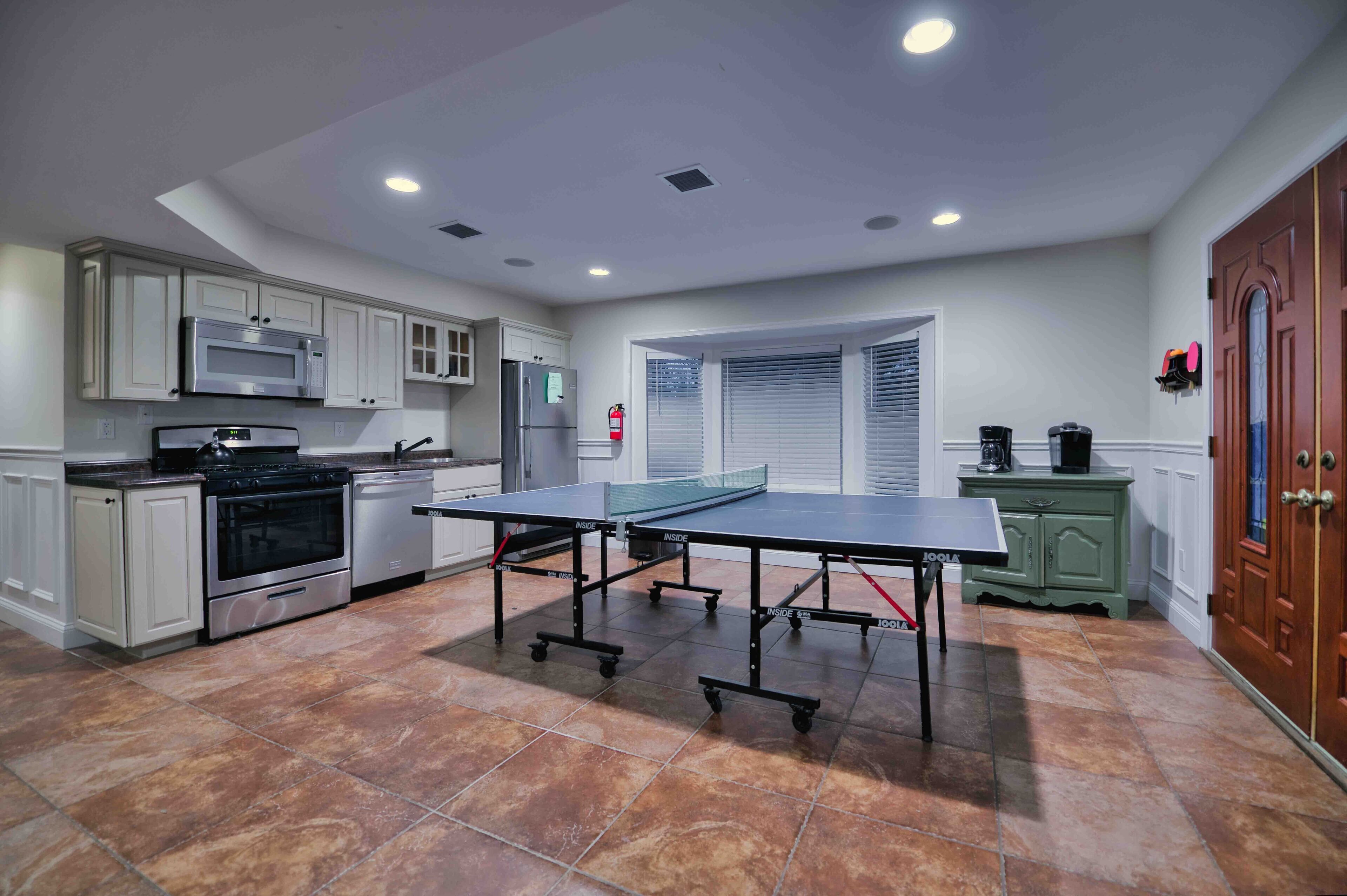 Una cocina con gabinetes blancos, electrodomésticos de acero inoxidable, una mesa de ping pong y un armario verde.