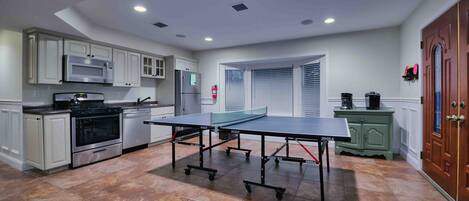 Una cocina con gabinetes blancos, electrodomésticos de acero inoxidable, una mesa de ping pong y un armario verde.