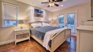 A bedroom with a bed, nightstands, a ceiling fan, a window with blinds, a door, and a view of the outside. - Beachside 8BR • 1 Block from Ocean (Surf City)