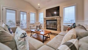 A living room with a stone fireplace, large windows, and a ceiling fan. - Beachside 8BR • 1 Block from Ocean (Surf City)
