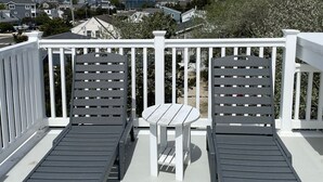 A balcony with two grey lounge chairs and a small white table, overlooking a residential area and a body of water under a partly cloudy sky. - Beachside 8BR • 1 Block from Ocean (Surf City)