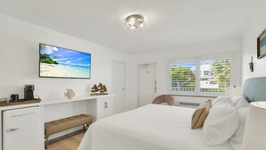 Quarto com cama, televisão, escrivaninha, um banquinho e janela com vista para o verde.
