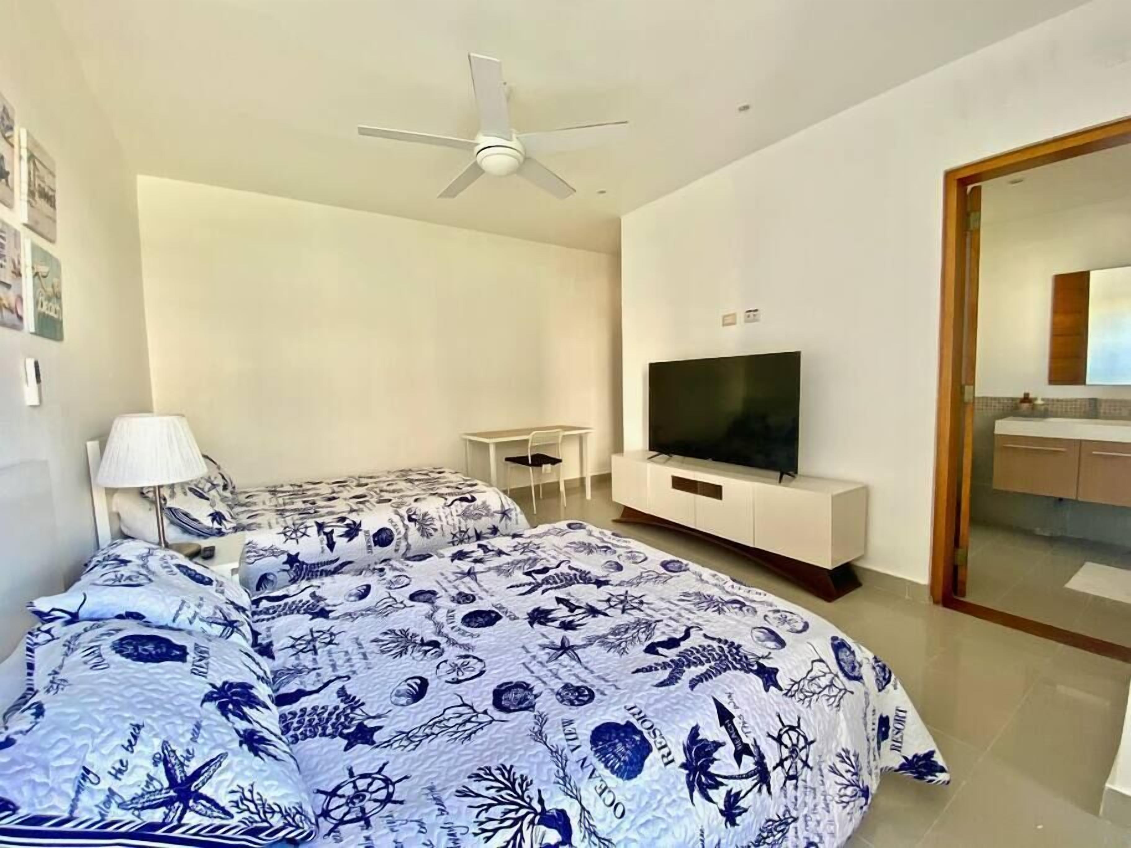 Une chambre à coucher moderne, avec un lit, une télévision, un ventilateur de plafond et une salle de bain visible à travers une porte ouverte.