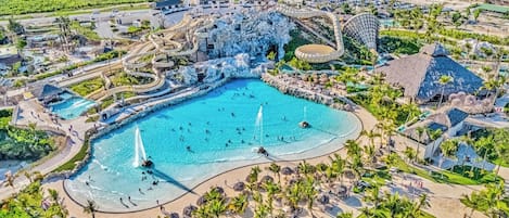 A large outdoor swimming pool with multiple water features, surrounded by palm trees and a tropical landscape.