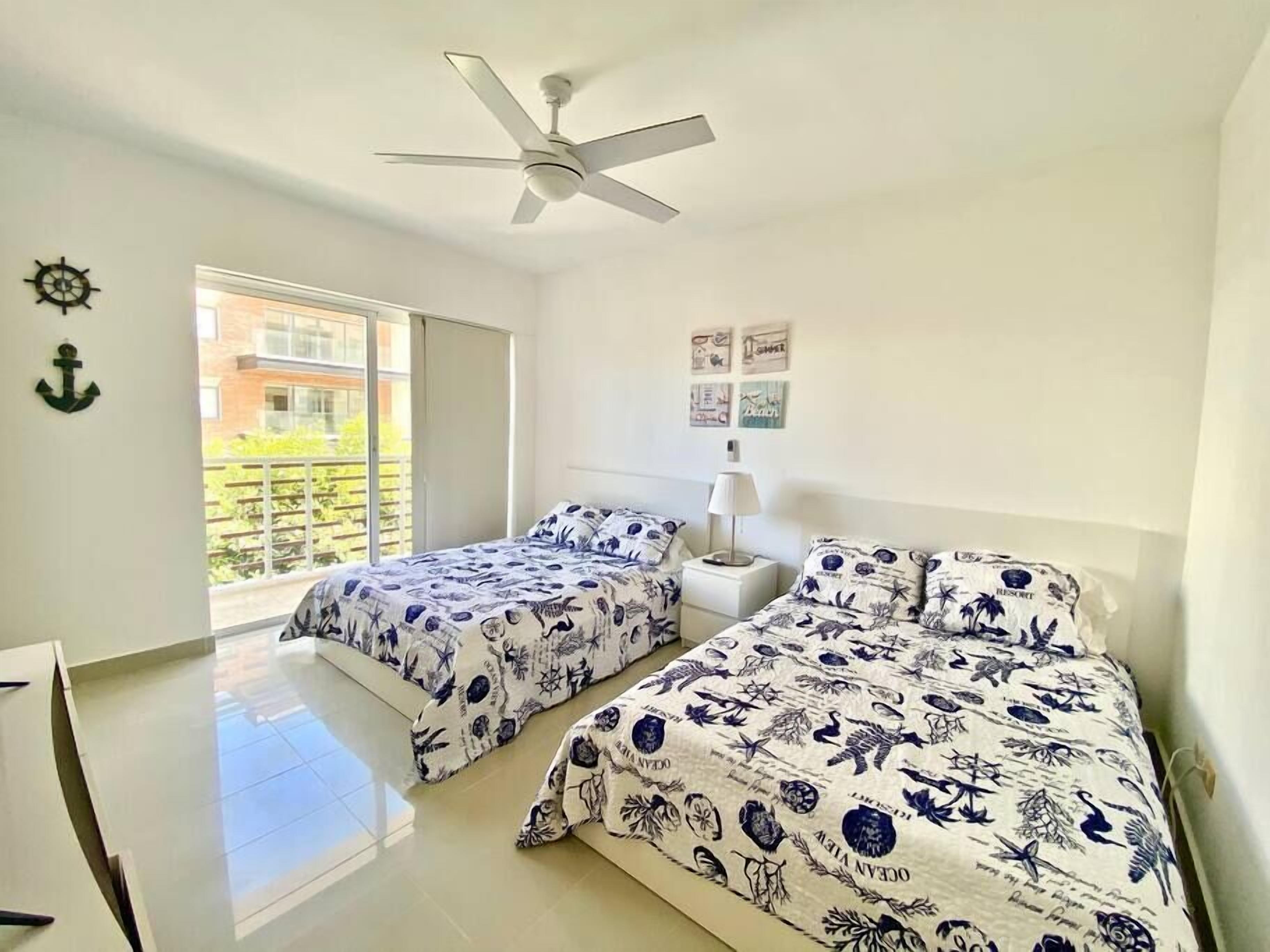 A bedroom with two beds, a ceiling fan, a window with a view of a building, and a door leading to a balcony.