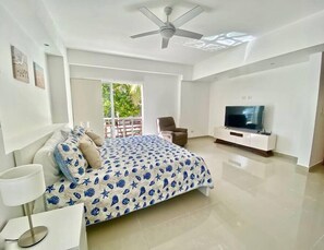 Un dormitorio moderno con cama, televisor, ventilador de techo y balcón con vistas a zonas verdes.