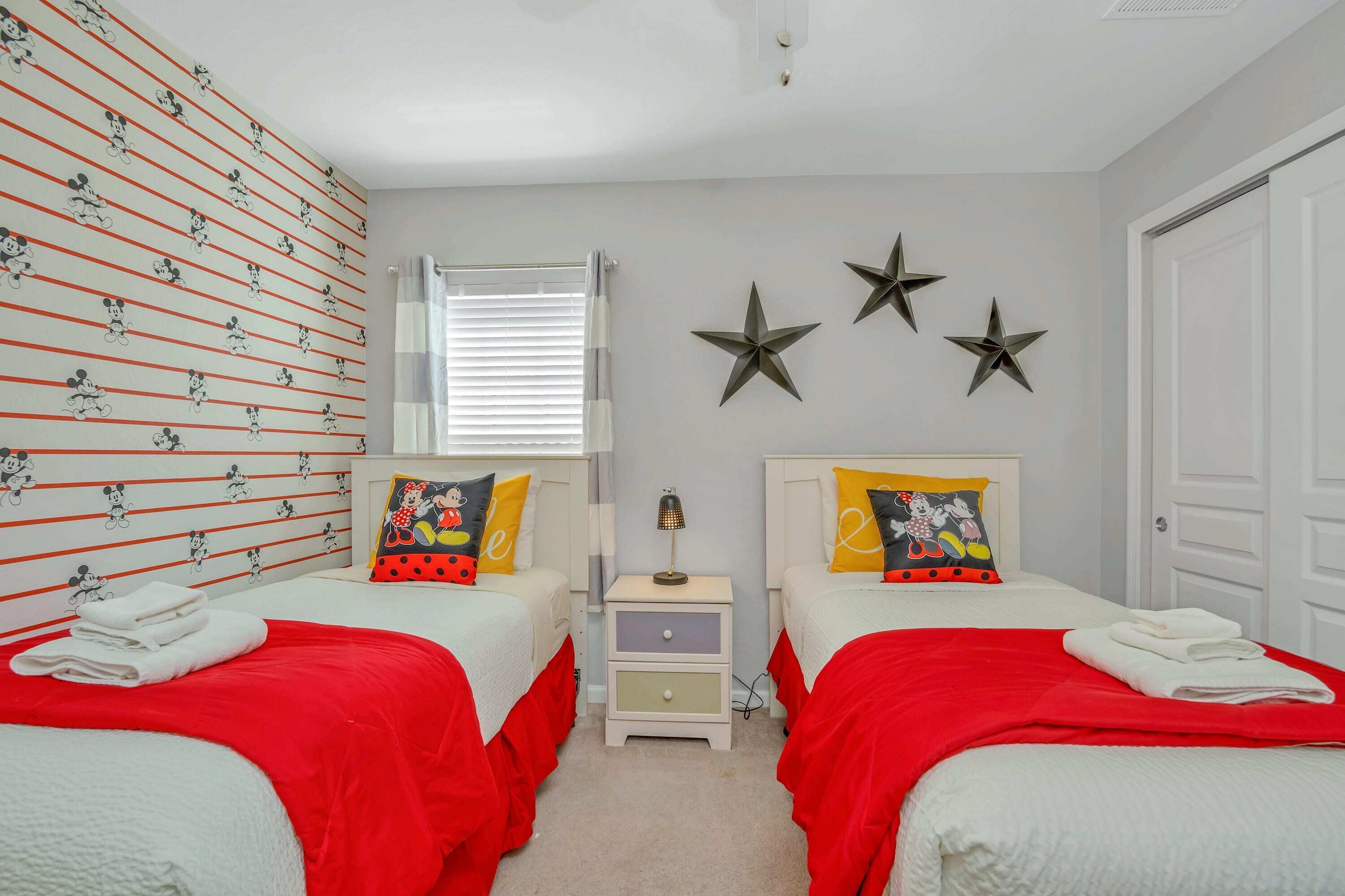 Une chambre avec deux lits, chacun recouvert d’une couverture rouge et agrémenté d’oreillers blancs, une table de chevet avec une lampe, et un mur recouvert d’un papier peint à l’effigie de Mickey Mouse.
