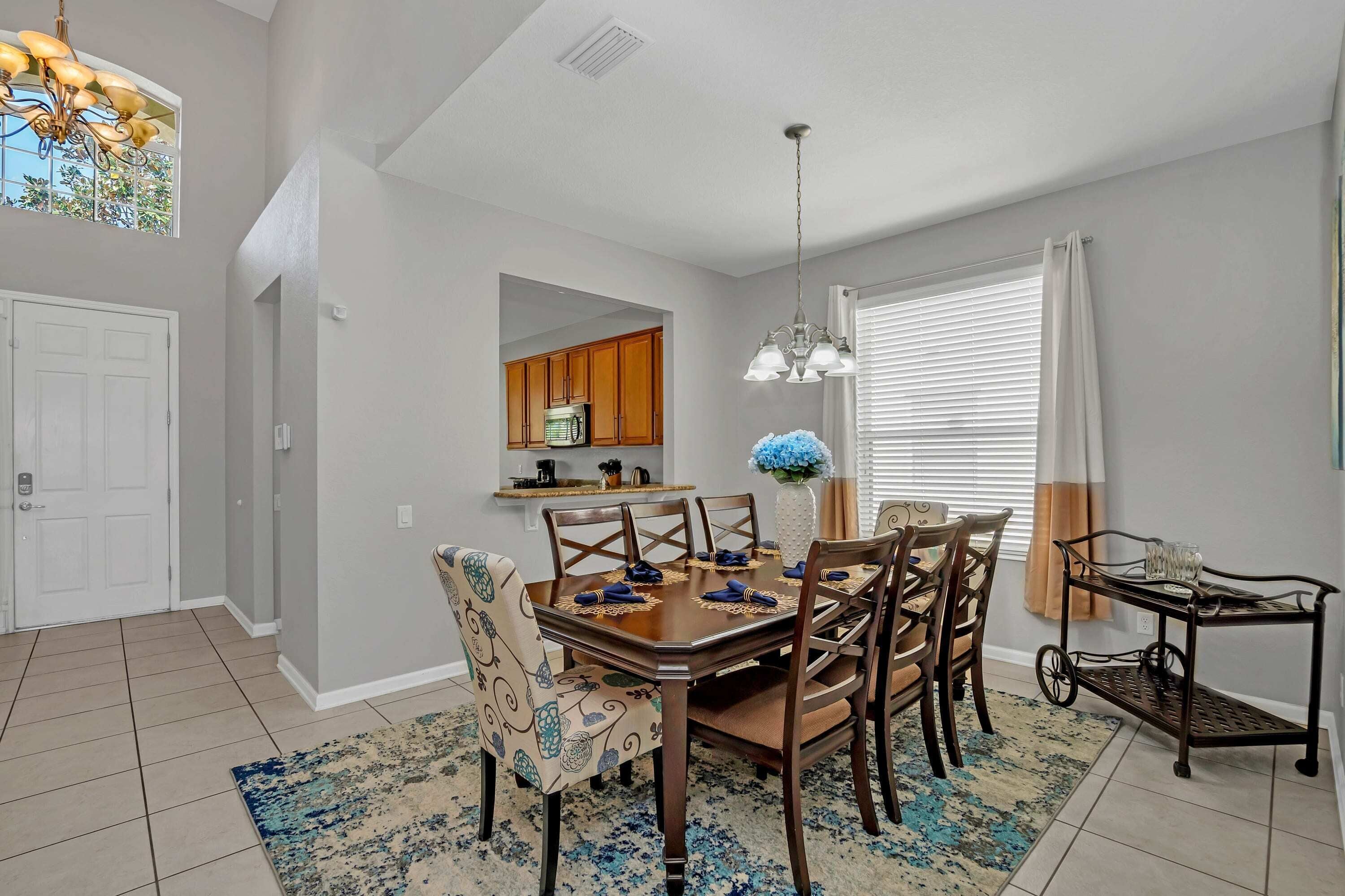 A dining area with a wooden table set for four, surrounded by patterned chairs. A chandelier hangs above, and a window with blinds is visible.
