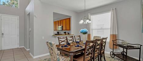 A dining area with a wooden table set for four, surrounded by patterned chairs. A chandelier hangs above, and a window with blinds is visible.