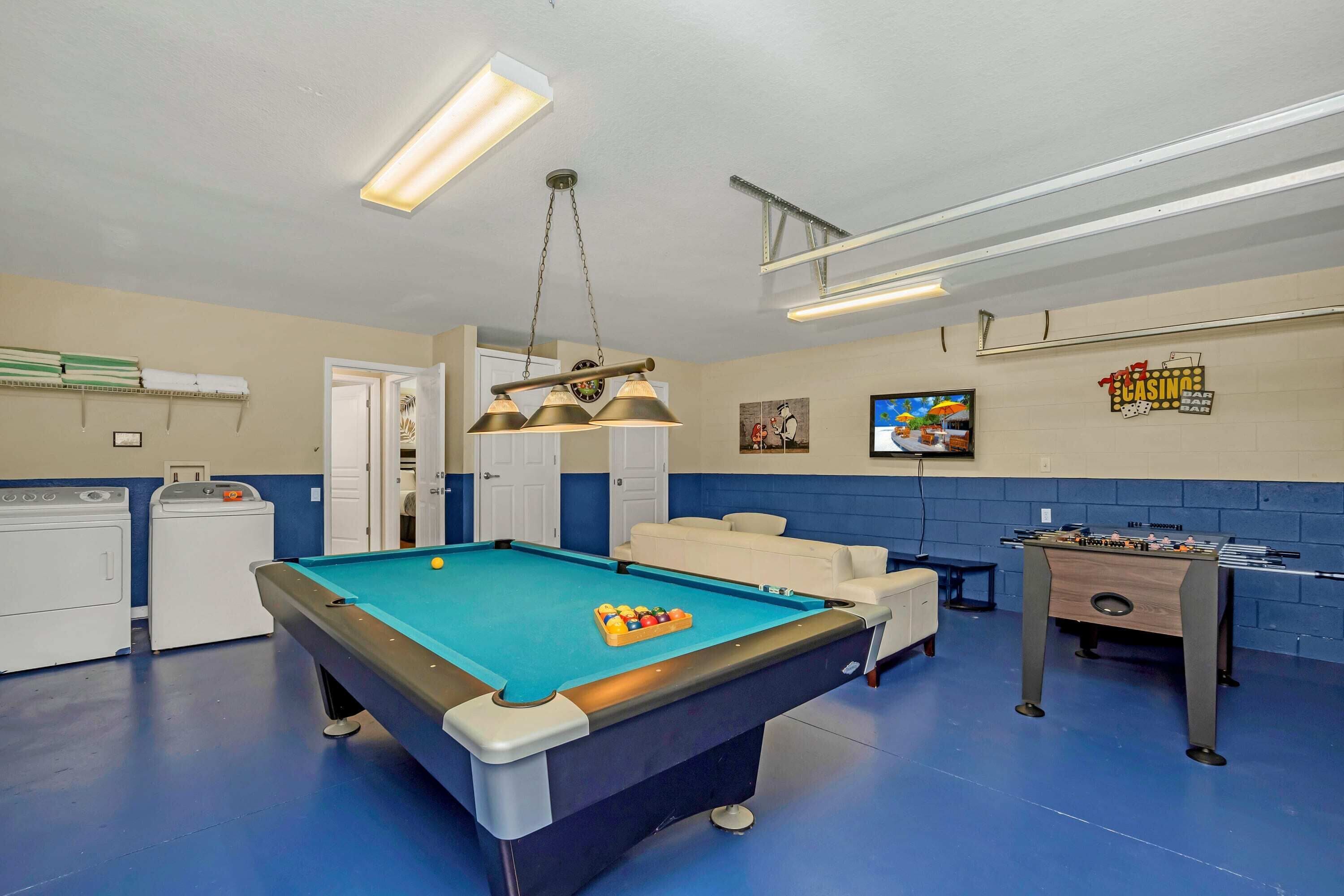 A pool table with a foosball table and a television in a room with blue walls and a ceiling fan.