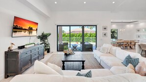 A spacious living room with a large sofa, a coffee table, and a painting of a sunset. There is a sliding glass door leading to an outdoor patio with a dining area.