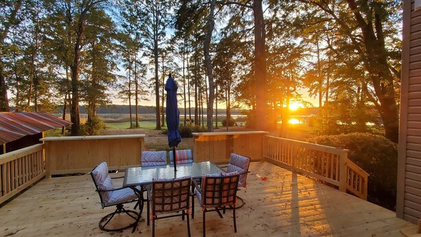 Eine Terrasse im Freien mit einem Tisch und Stühlen mit Glastischplatte, umgeben von Holzgeländern und Blick auf Bäume und einen Sonnenuntergang.