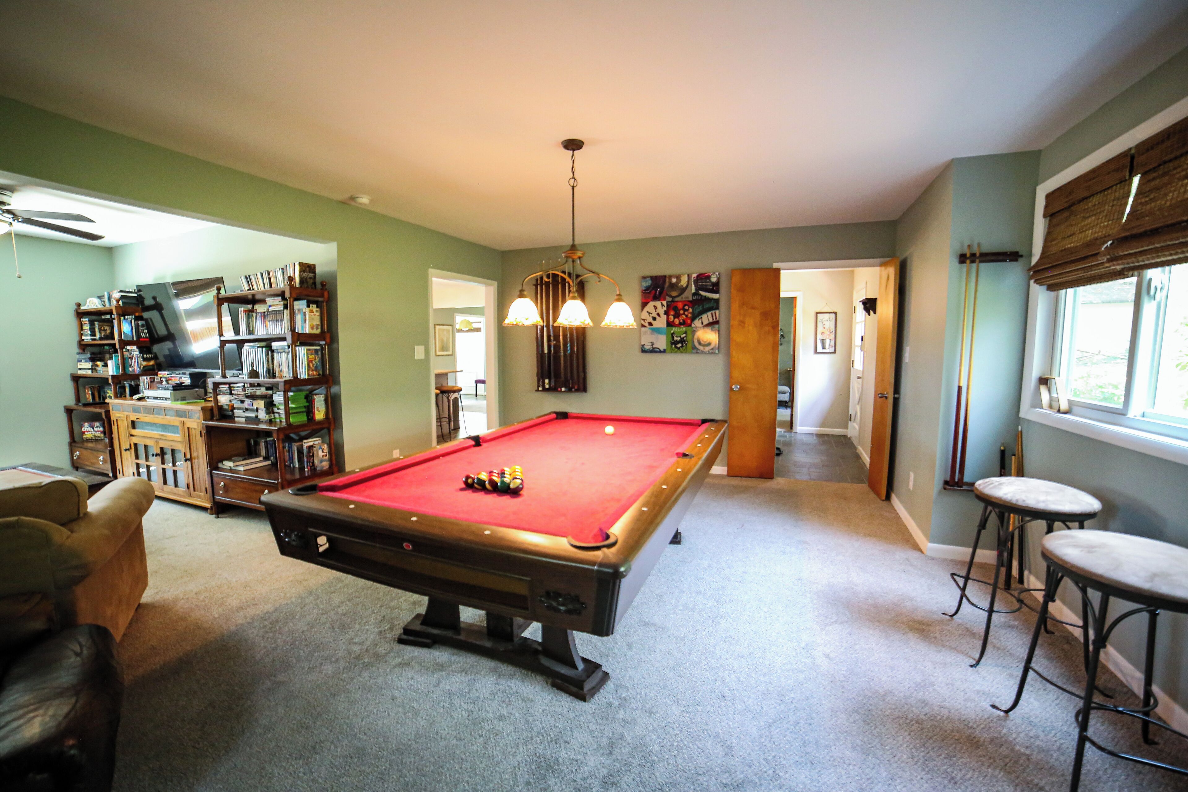 A pool table in a room with a ceiling fan, a bookshelf, and a window with blinds.