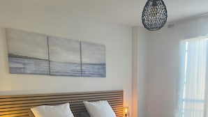 Ein ordentlich bezogenes Bett mit weißen Leinen, ein Teppich mit grünem Muster, ein an der Wand hängendes Kunstwerk und eine Pendelleuchte.