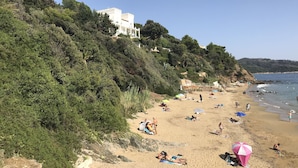 Ein Strand mit badenden und sonnenden Menschen, umgeben von viel Grün und einem weißen Gebäude.