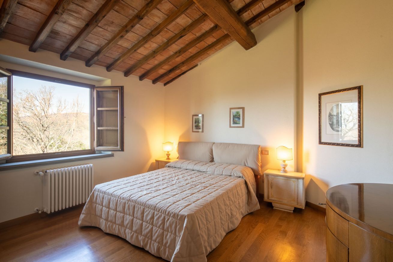 Une chambre avec un plafond en bois, un lit recouvert d’une courtepointe, des tables de chevet avec des lampes, un radiateur et une fenêtre donnant sur un beau paysage.