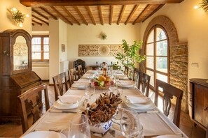 A dining room with a long table set for a meal, wooden chairs, a stone wall, and a large arched window.