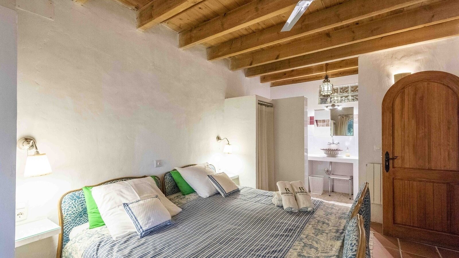 A bedroom with a wooden ceiling, a bed with striped bedding, a nightstand, a wall-mounted lamp, and a wooden door.