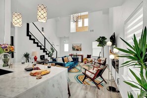 A spacious living area with a staircase, a blue sofa, a dining table with a fruit platter, and modern lighting fixtures. - Hottub-Deck-Firepit-Cornhole! (Fredericksburg)