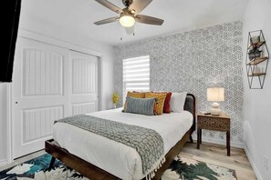 Un dormitorio con cama, ventilador de techo, ventana con persianas, mesita de noche y pared con diseño.
