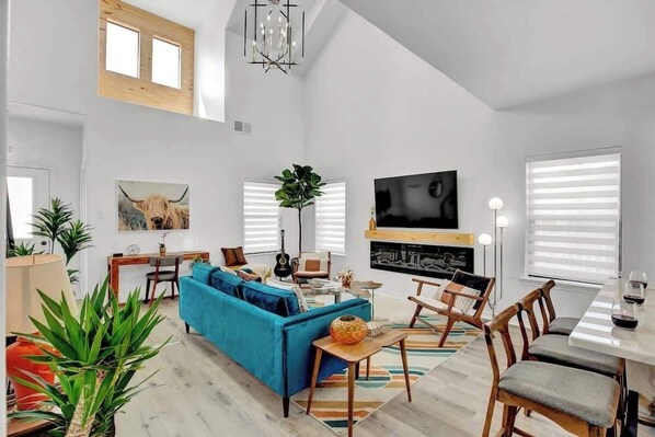 Amplia sala de estar con un sofá azul, sillas de madera, una mesa de comedor con sillas, un televisor montado en la pared y un gran ventanal con persianas.