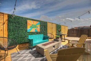 An outdoor patio with a green wall, a turquoise sofa, wooden chairs, and a fire pit.