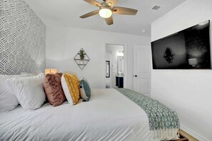 Un dormitorio con cama, almohadas, ventilador de techo, televisor montado en la pared y un diseño geométrico en la pared.