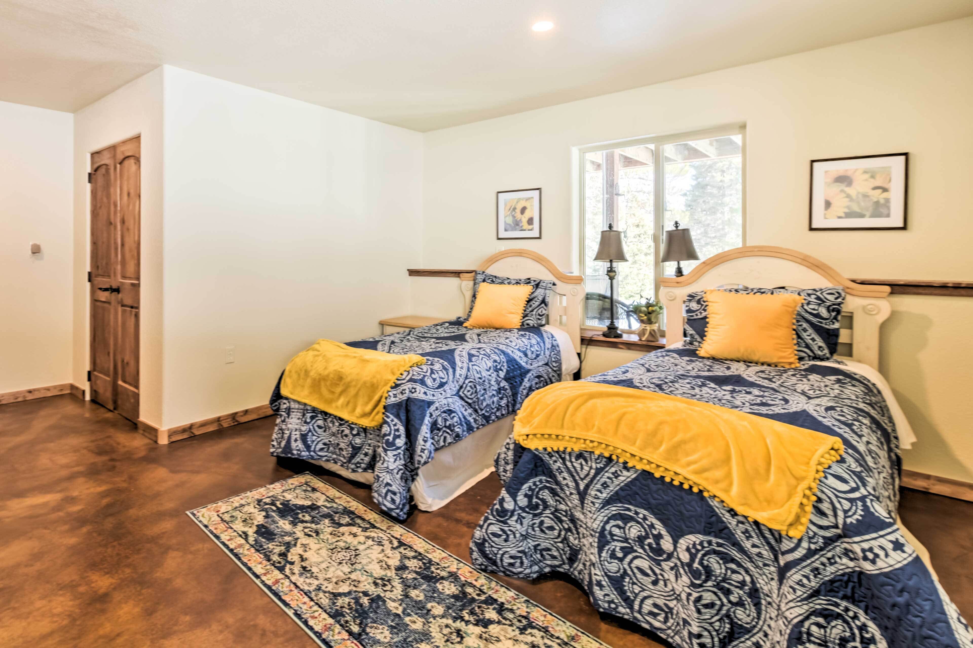 A bedroom with two beds, each with a blue and white patterned bedspread and a yellow throw blanket. There is a window with a view of greenery outside, a wooden door, and a small table with a lamp.