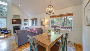 Un vaste salon comprenant une table à manger en bois, une cheminée, un téléviseur et de grandes fenêtres donnant sur l’extérieur.