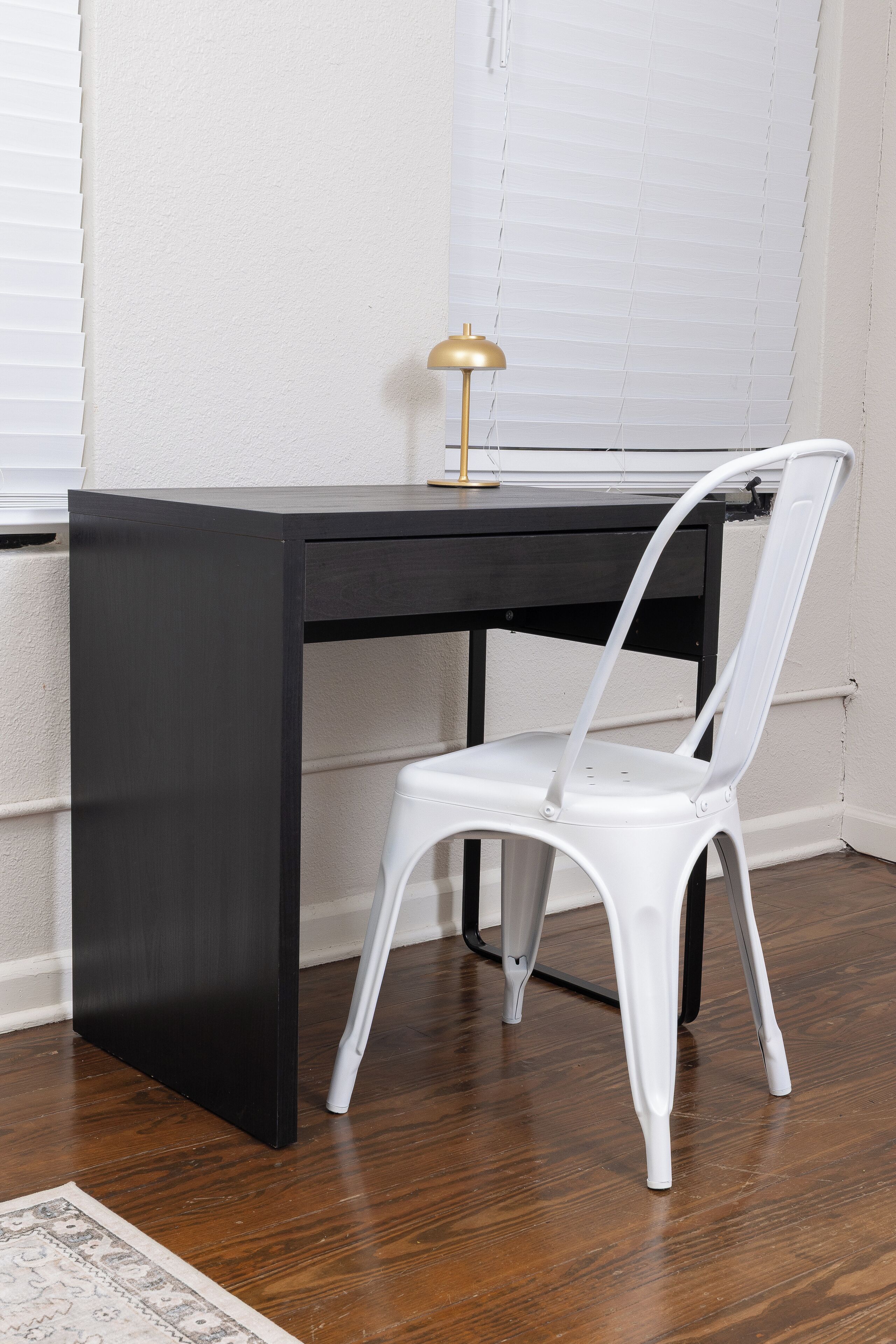 A dark wooden desk with a white chair, a gold lamp, and a patterned rug on a wooden floor.